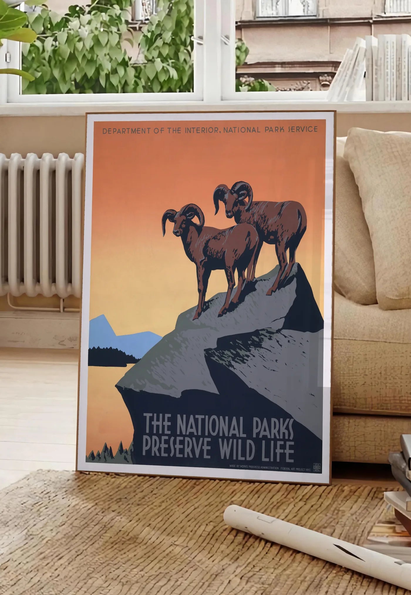 Poster affiche mouflons National Parks vintage, posee au sol dans un salon parisien, devant une grande fenetre donnant sur un immeuble haussmannien, entre un ficus et un canape creme